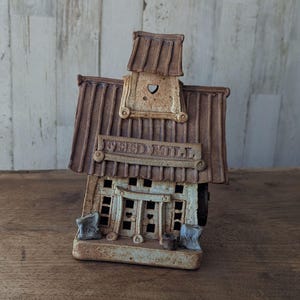 May include: A ceramic feed mill sculpture featuring a brown textured roof with a small heart cutout in the gable. The off-white facade displays the embossed text FEED MILL above a central doorway with small windows and two small sacks at the base.