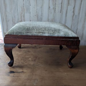 May include: An antique footstool with a rectangular, light green upholstered cushion. The dark brown wooden frame features curved legs. The footstool is placed on a wooden surface, showcasing its vintage design and craftsmanship.