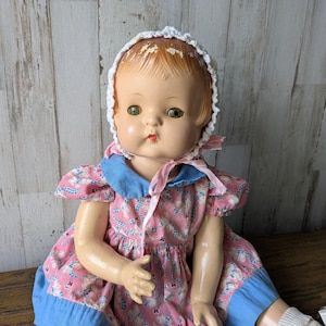 Puede incluir: Una muñeca de porcelana vintage con un vestido rosa y azul y un gorro blanco de ganchillo. La muñeca tiene ojos verdes y una sonrisa pintada.