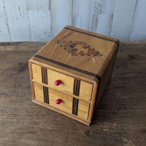 Op de afbeelding: Een kleine houten ladekast met twee lades, voorzien van inlegwerk van licht en donker hout. Het bovenoppervlak toont een ruitvormig inlegwerk met een hondensilhouet. Elke lade is voorzien van een kleine, ronde, felrode knop als handgreep.