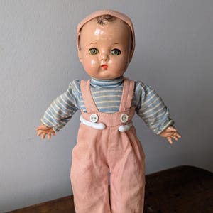 Puede incluir: Una muñeca de porcelana vintage con ojos verdes pintados, mejillas sonrosadas y cabello castaño. La muñeca lleva una camisa a rayas rosa y azul y un mono rosa con botones blancos.