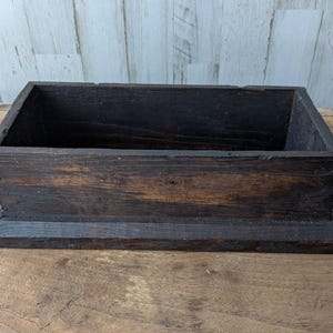 May include: A rectangular, dark brown wooden box with a flat base and visible wood grain. The box features straight sides and a hollow interior, constructed with visible joinery details along the corners and edges of the dark stained wood surface.