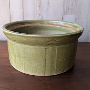 Puede incluir: Una olla redonda de cerámica verde claro con un borde ancho y líneas verticales. El borde tiene un patrón decorativo de pequeños chevrones. El interior tiene una banda marrón rojiza más oscura cerca de la parte superior. La olla está sobre una superficie de madera oscura.