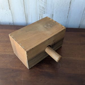 May include: A rectangular wooden mallet with a cylindrical handle. The mallet is constructed from light brown wood, with visible joinery at the corners. The handle is attached to the side of the mallet.