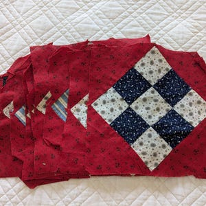 May include: A stack of red fabric quilt squares with a floral pattern. Some squares feature a diamond pattern with blue and white squares. The quilt squares are arranged on a white quilted surface.