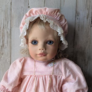 May include: A porcelain doll with blue eyes and blonde hair, wearing a pink gingham dress and matching bonnet trimmed with white lace. A gold-colored necklace is visible. The doll's face is painted with detailed features.