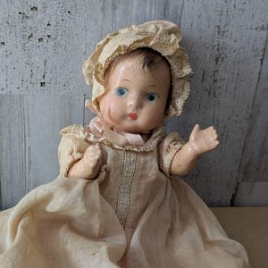 May include: A vintage doll with a cracked face, blue eyes, and painted red lips. The doll wears a cream-colored lace bonnet and a matching long-sleeved gown with a central vertical lace panel and a ruffled collar, featuring a light pink ribbon tie.