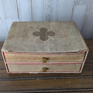 May include: A vintage beige jewelry box with two drawers. The top features an embossed floral design in brown. The drawers have decorative trim with red accents and small brass-colored handles. The box sits on a wooden surface.