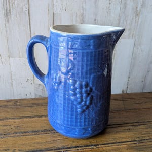 May include: A blue ceramic pitcher with a handle and a spout. The pitcher has a textured surface with a grape design. The interior is white. The pitcher is sitting on a wooden surface.