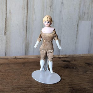 May include: A vintage porcelain doll with a blonde bob hairstyle, blue eyes, and painted facial features. The doll wears a tattered brown fabric outfit with white porcelain arms and legs. It stands on a white metal stand, showcasing its antique charm.