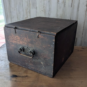 Puede incluir: Una caja de almacenamiento rectangular de madera con un acabado marrón oscuro desgastado. La caja cuenta con una tapa con bisagras asegurada por dos pequeños pestillos metálicos y un asa central con placa decorativa.