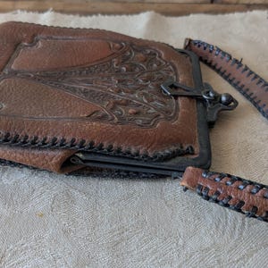 May include: A vintage brown leather wallet with a decorative embossed design on the front. The wallet has a metal clasp closure and a matching leather strap. The edges are stitched with black thread, and the wallet is resting on a cream-colored cloth.