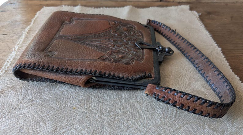 May include: A vintage brown leather wallet with a decorative embossed design on the front. The wallet has a metal clasp closure and a matching leather strap. The edges are stitched with black thread, and the wallet is resting on a cream-colored cloth.