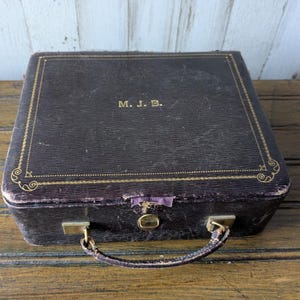 May include: A vintage, rectangular, dark brown case with gold trim and the initials "M. J. B." on the top. The case has a handle, a clasp, and metal hardware. The case appears to be made of leather or a similar material.