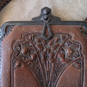 May include: A brown leather purse with a decorative floral design and a metal clasp. The purse has a stitched edge and a brown leather strap. The clasp is silver-colored. The purse is rectangular in shape.
