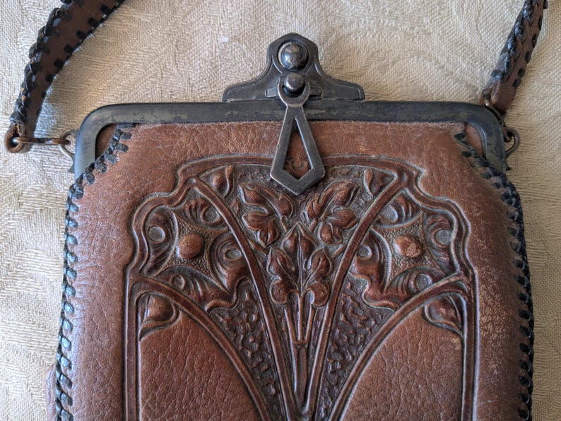 May include: A brown leather purse with a decorative floral design and a metal clasp. The purse has a stitched edge and a brown leather strap. The clasp is silver-colored. The purse is rectangular in shape.