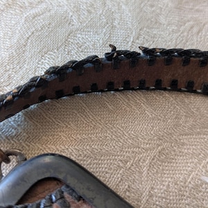 May include: Close-up of a brown leather belt with a braided edge and a metal buckle. The belt has a worn appearance, with visible stitching and a metal clasp. The leather is a medium brown color.