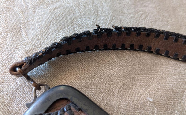 May include: Close-up of a brown leather belt with a braided edge and a metal buckle. The belt has a worn appearance, with visible stitching and a metal clasp. The leather is a medium brown color.