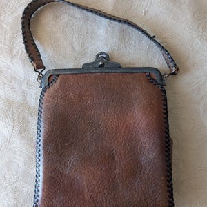 May include: A brown leather handbag with a textured surface and a stitched border. The bag features a metal clasp and a matching leather handle. The bag measures approximately 7 inches in height and 5 inches in width.