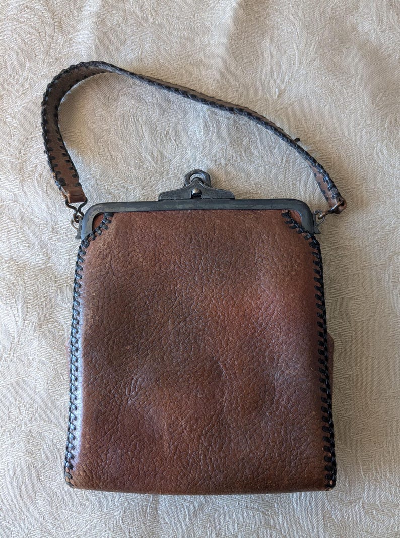 May include: A brown leather handbag with a textured surface and a stitched border. The bag features a metal clasp and a matching leather handle. The bag measures approximately 7 inches in height and 5 inches in width.