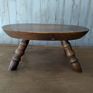 May include: A small, oval-shaped wooden stool with a smooth, dark brown top surface and three turned legs. The legs feature a rounded, tapered design that flares outward, providing a stable base for the low-profile, solid wood seat construction.