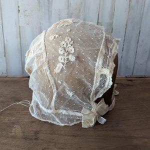 May include: An off-white lace bonnet featuring a delicate Swiss dot pattern and intricate floral embroidery. The headwear is constructed with sheer fabric panels, scalloped edges, and a small fabric bow detail positioned at the side near the base.
