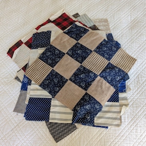 Può includere: Una collezione di quadrati di tessuto patchwork in vari modelli e colori. I quadrati presentano disegni a quadretti, floreali e a righe in tonalità di blu, marrone, rosso e bianco. I tessuti sono disposti su una superficie trapuntata bianca.
