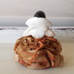 May include: A small porcelain figurine of a woman wearing a white dress and a black hat. The figurine is sitting on a fabric cushion with a gold and orange pattern.