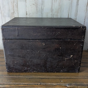 May include: A dark brown, rectangular trunk with a textured surface. The trunk is made of a sturdy material and has a hinged lid. It sits on a wooden surface, with a light-colored wooden wall in the background.