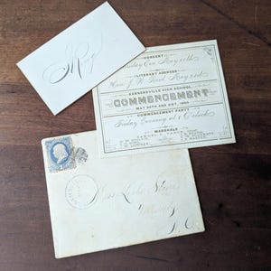 May include: A collection of antique paper items: a Kernersville High School commencement invitation from May 20th and 21st, 1886, a card with the initials "S. Ring", and an envelope with a blue stamp. The items are on a wooden surface.