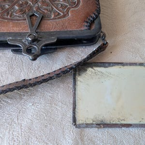 May include: An antique brown leather purse with a decorative floral design and a metal clasp. A small rectangular mirror with a metal frame is next to the purse. The purse has a long, thin strap.