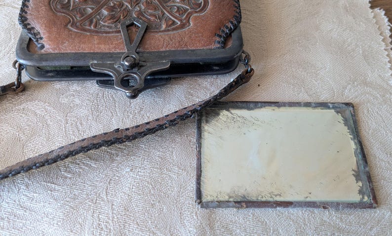 May include: An antique brown leather purse with a decorative floral design and a metal clasp. A small rectangular mirror with a metal frame is next to the purse. The purse has a long, thin strap.