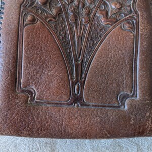 May include: Close-up of a brown leather wallet with an embossed Art Nouveau design. The wallet features a central floral motif and decorative border. The edges are stitched with black thread, and the leather has a textured appearance.