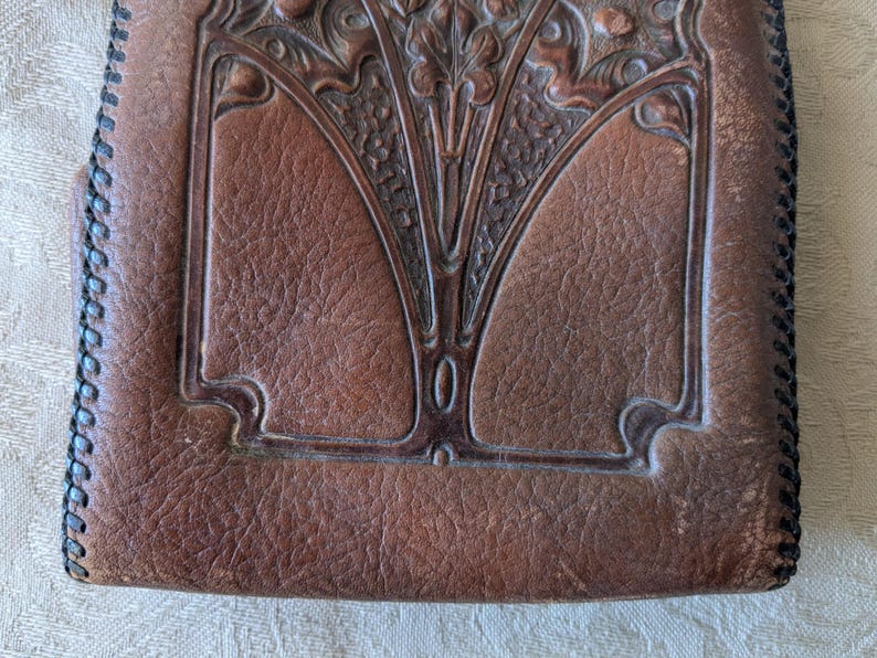 May include: Close-up of a brown leather wallet with an embossed Art Nouveau design. The wallet features a central floral motif and decorative border. The edges are stitched with black thread, and the leather has a textured appearance.