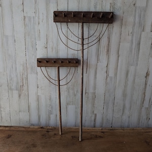 Op de afbeelding: Twee bruine houten tuinharken met lange stelen en metalen tanden. Elke hark heeft een horizontale houten kop met meerdere pinnen en een reeks gebogen metalen draden die een halfronde ondersteuningsstructuur onder de kop vormen.
