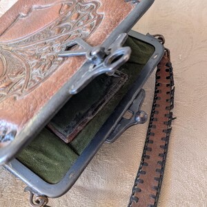 May include: An open, vintage brown leather purse with a decorative embossed design on the top flap. The purse has a silver metal frame and clasp, with a dark green interior and a brown leather strap.