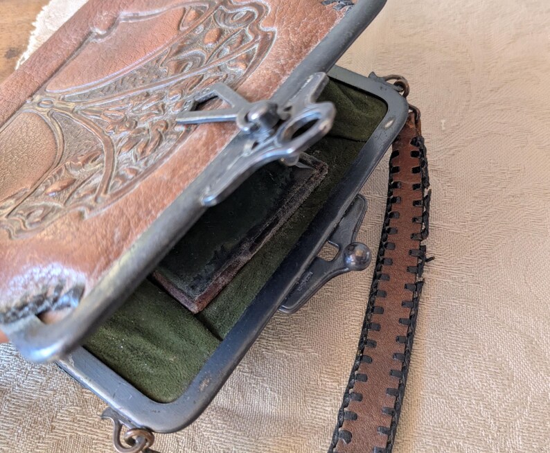 May include: An open, vintage brown leather purse with a decorative embossed design on the top flap. The purse has a silver metal frame and clasp, with a dark green interior and a brown leather strap.