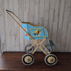 Può includere: Passeggino giocattolo vintage blu e oro con illustrazioni di un gatto e un clown. Il passeggino ha quattro ruote con dettagli dorati e un telaio pieghevole. Il manico è dorato e il sedile è azzurro.
