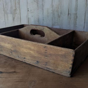 Könnte beinhalten: Ein rustikaler Holzorganizer mit Griff und mehreren Fächern. Die rechteckige Box besteht aus gealtertem Holz mit sichtbarer Maserung und Knoten, das ein natürliches, verwittertes Aussehen aufweist. Die Fächer sind durch Holzunterteilungen geteilt.