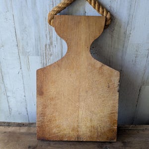 May include: A vintage wooden cutting board with a rope handle. The board is rectangular with a unique curved top and shows signs of wear, including scratches and discoloration, giving it a rustic appearance. The rope handle is made of twisted natural fibers.