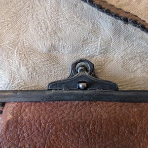 May include: Close-up of a brown leather purse with a metal clasp. The purse has a textured leather surface and a dark metal frame. The strap is visible at the top, and the clasp is in the center.