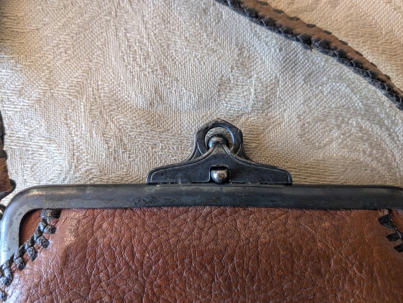 May include: Close-up of a brown leather purse with a metal clasp. The purse has a textured leather surface and a dark metal frame. The strap is visible at the top, and the clasp is in the center.