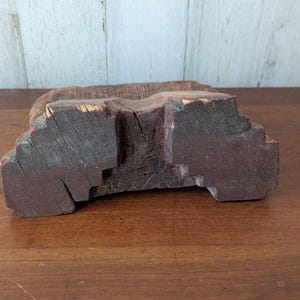Puede incluir: Un bloque de madera tallada y envejecida, de color marrón rojizo oscuro. El bloque tiene una superficie rugosa y texturizada con vetas visibles y una sección recortada. La madera parece envejecida y posiblemente reutilizada.