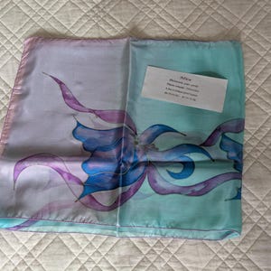 May include: A square silk scarf featuring a hand-painted floral design in shades of purple, blue, and turquoise. The scarf has a light pink border and includes a white rectangular card with the text Mira Peinte sur soie, Pièce unique, Décoration, 6 Rue le Chanoine, 06110 Le Cannet, 04 93 38 25 45 - 06 10 70 72 08.