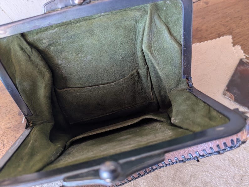 May include: Open view of a vintage brown leather purse with a metal clasp. The interior is lined with olive green fabric and features a small pocket. The purse is open, revealing its contents.
