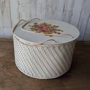 Może przedstawiać: Okrągły, kremowy kosz z tkaniny z dopasowaną pokrywą. Pokrywa ma kwiatowy wzór w odcieniach czerwieni, różu i zieleni. Dołączony jest sznurkowy uchwyt. Kosz nadaje się do przechowywania, o średnicy około 30 cm.