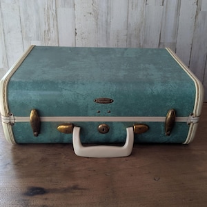May include: A rectangular suitcase with a textured teal exterior and cream-colored trim. The case features a central cream handle, a central keyhole, and two brass-toned metal latches on the front. The surface has a mottled, weathered appearance.