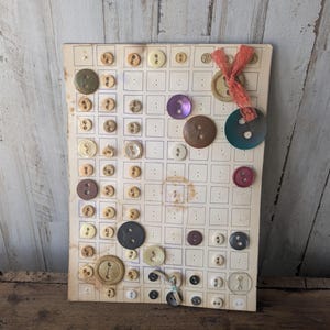 May include: A vintage button card featuring an assortment of buttons in various sizes, shapes, and colors. The buttons are arranged on a grid, with colors including brown, cream, purple, and black. A decorative ribbon is attached to the top.