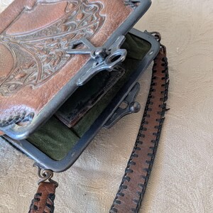 May include: Vintage brown leather purse with a decorative embossed design on the front. The purse has a metal clasp closure and a matching leather strap with stitched detailing. The interior is lined with green fabric.