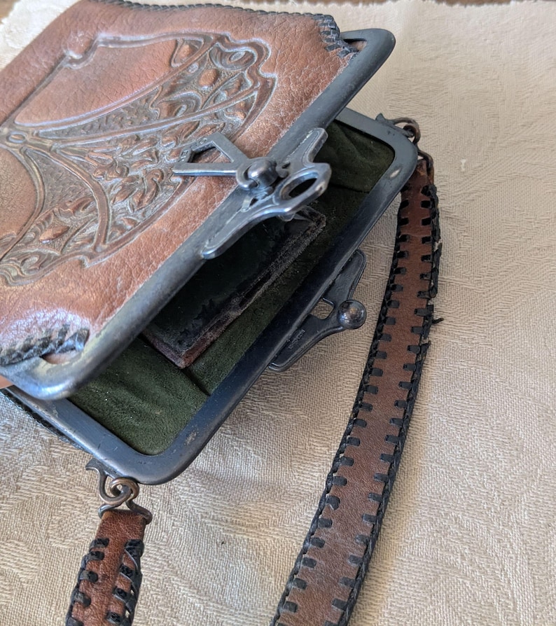 May include: Vintage brown leather purse with a decorative embossed design on the front. The purse has a metal clasp closure and a matching leather strap with stitched detailing. The interior is lined with green fabric.